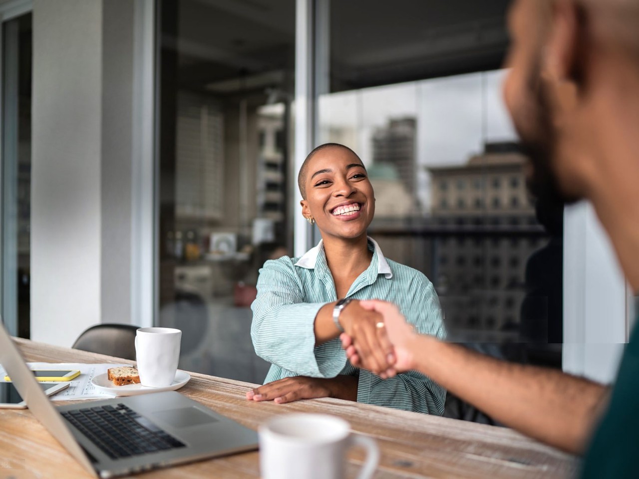 A smiling woman shakes hands with an advisor across a table with coffee and a laptop