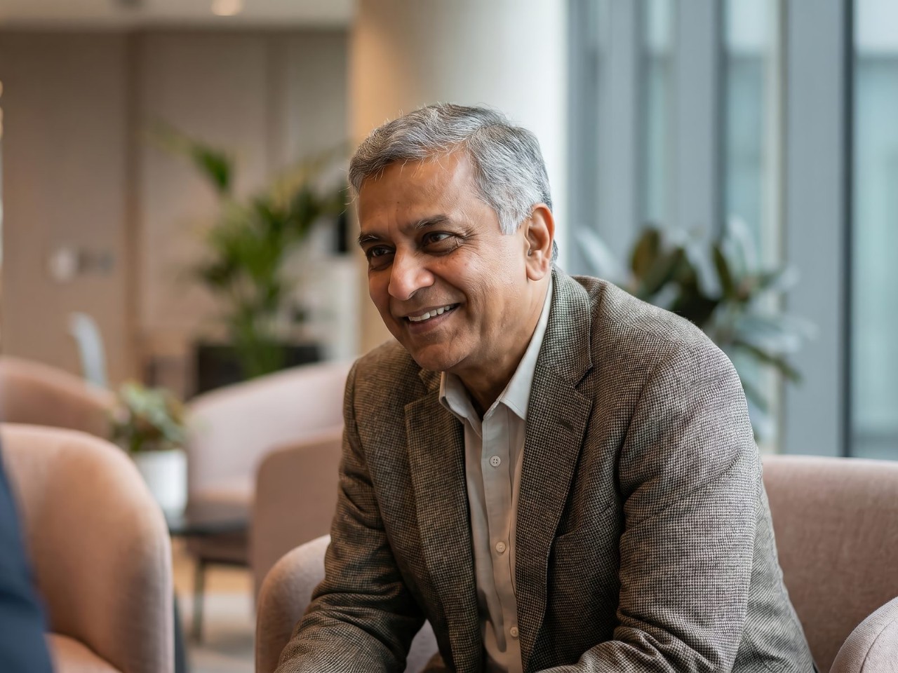 A smiling senior advisor in a tweed jacket sits in a bright office lounge