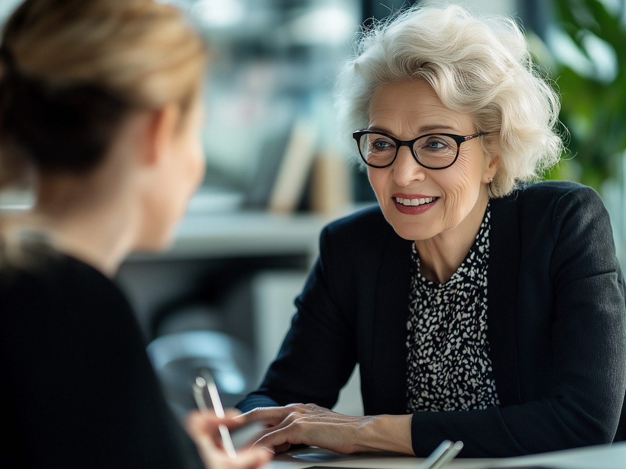 An advisor in glasses smiles as she listens to a client across a desk